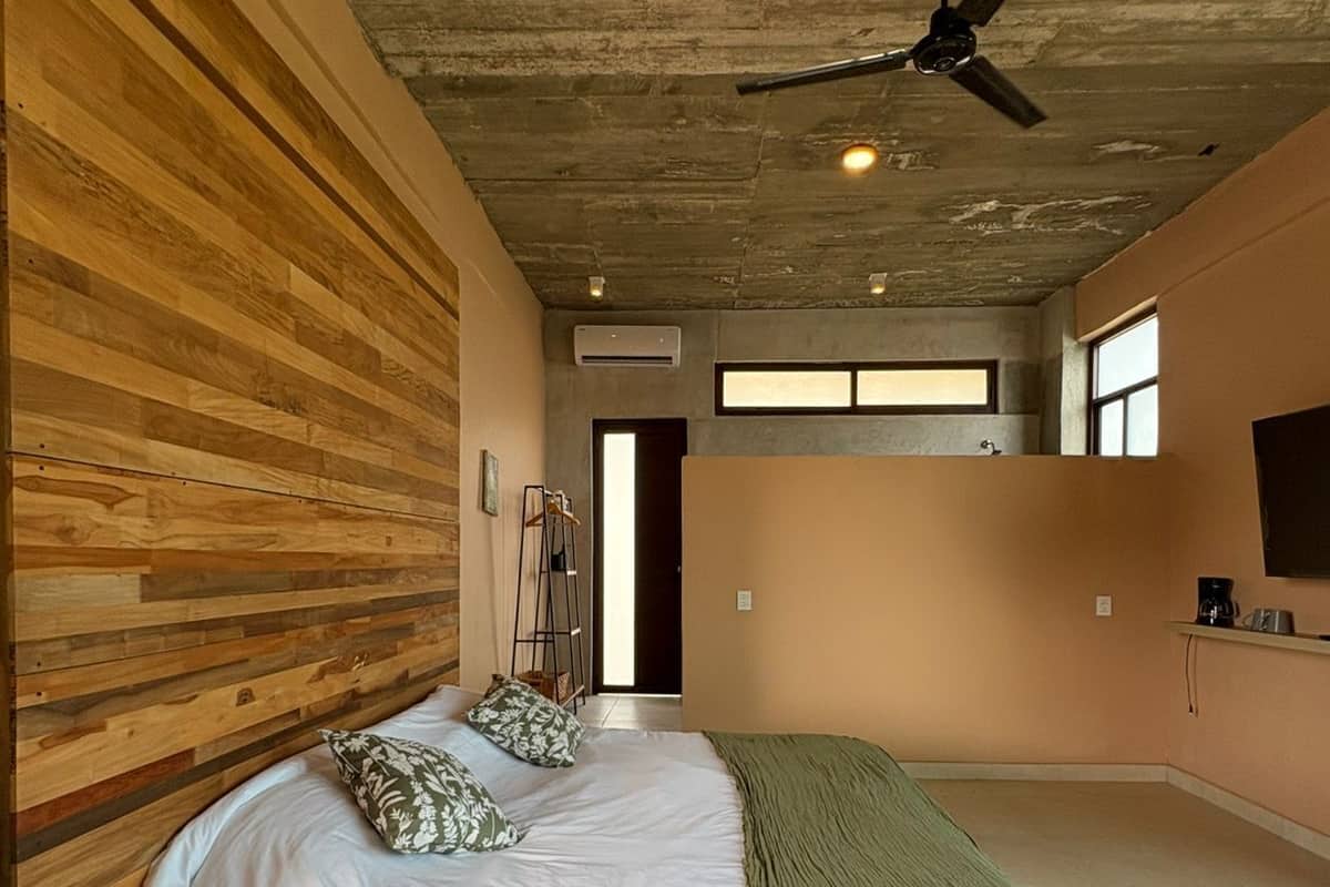 Une chambre avec une tête de lit en bois, un lit avec une literie blanche et un plaid vert, un ventilateur de plafond et une vue sur la cuisine.
