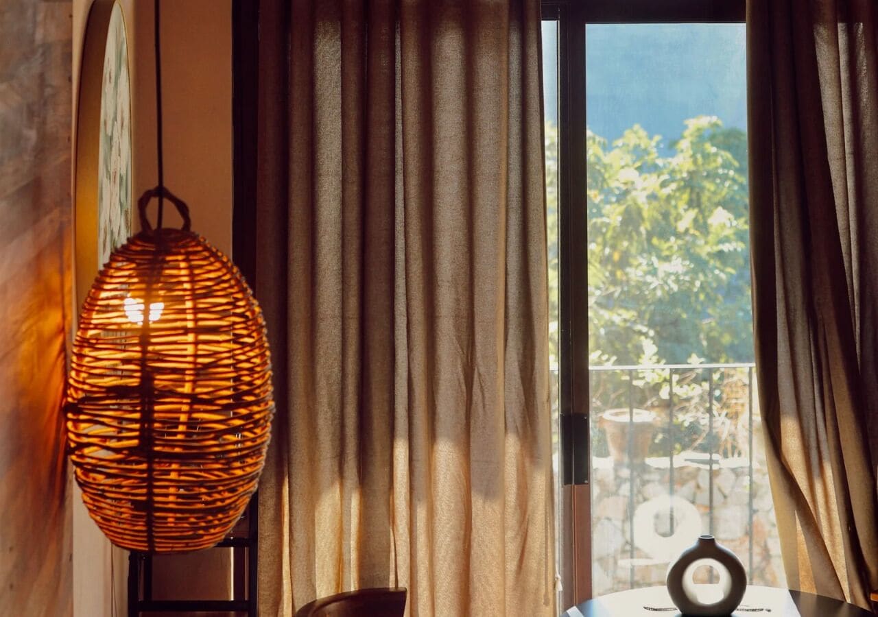 Una habitación con una lámpara de mimbre, cortinas y vistas a un balcón con follaje.
