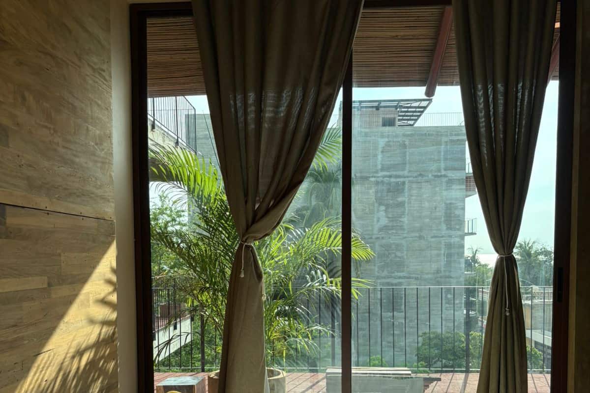 Une chambre à coucher avec un lit, des rideaux et une vue sur un balcon agrémenté de plantes et donnant sur un immeuble.
