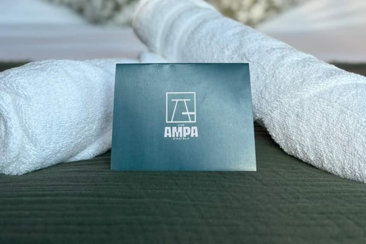 Un lit bien fait, avec une serviette blanche pliée et une carte portant l’inscription « AMPA », le tout posé sur un plancher en bois.
