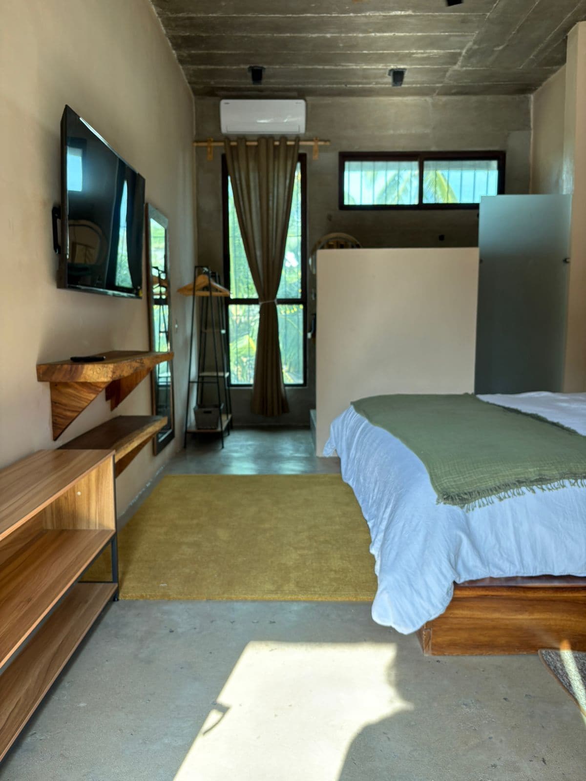A bedroom with a bed, a TV mounted on the wall, a wooden bench, and a window with curtains.