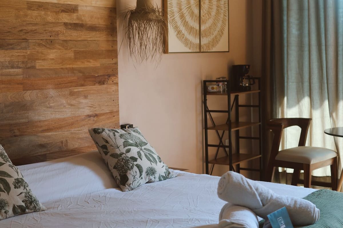 Une chambre à coucher avec une tête de lit en bois, un lit agrémenté d’oreillers à motifs floraux, un luminaire suspendu, une machine à café et une chaise.