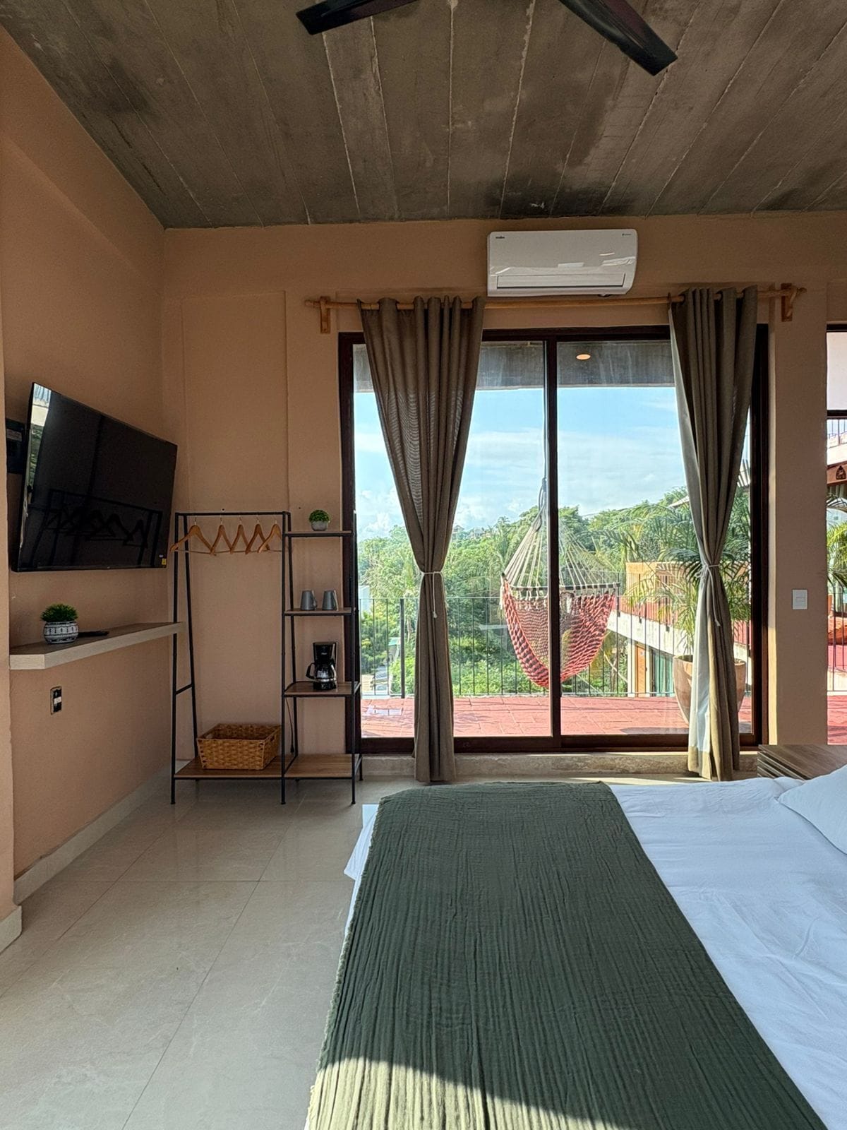 A hotel room with a bed, a TV, a shelf with items, and a view of the outdoors.