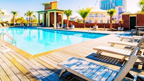 A swimming pool with wooden deck and lounge chairs, surrounded by palm trees and a brick wall. - Tannin Luxe 3BR + Loft Bunks, Steps To Beach Access, Pool, Firepit, Water Views (Orange Beach)