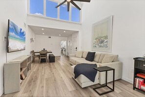 A spacious living area with a high ceiling, a dining table, a sofa, and a bookshelf.