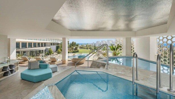 A modern indoor pool area with a clear view of the outside, a seating area with cushions, and a pool with a textured ceiling.