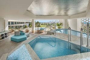 A modern indoor pool area with a clear view of the outside, a seating area with cushions, and a pool with a textured ceiling.