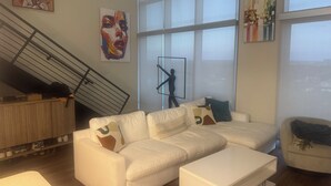 A modern living room with a white sectional sofa, a white coffee table, and a staircase with a railing. There are abstract paintings on the walls and a large window providing natural light. - Luxury Loft Penthouse in the Heart of the City with Skyline Views – 1BR, 1.5BA (Austin)