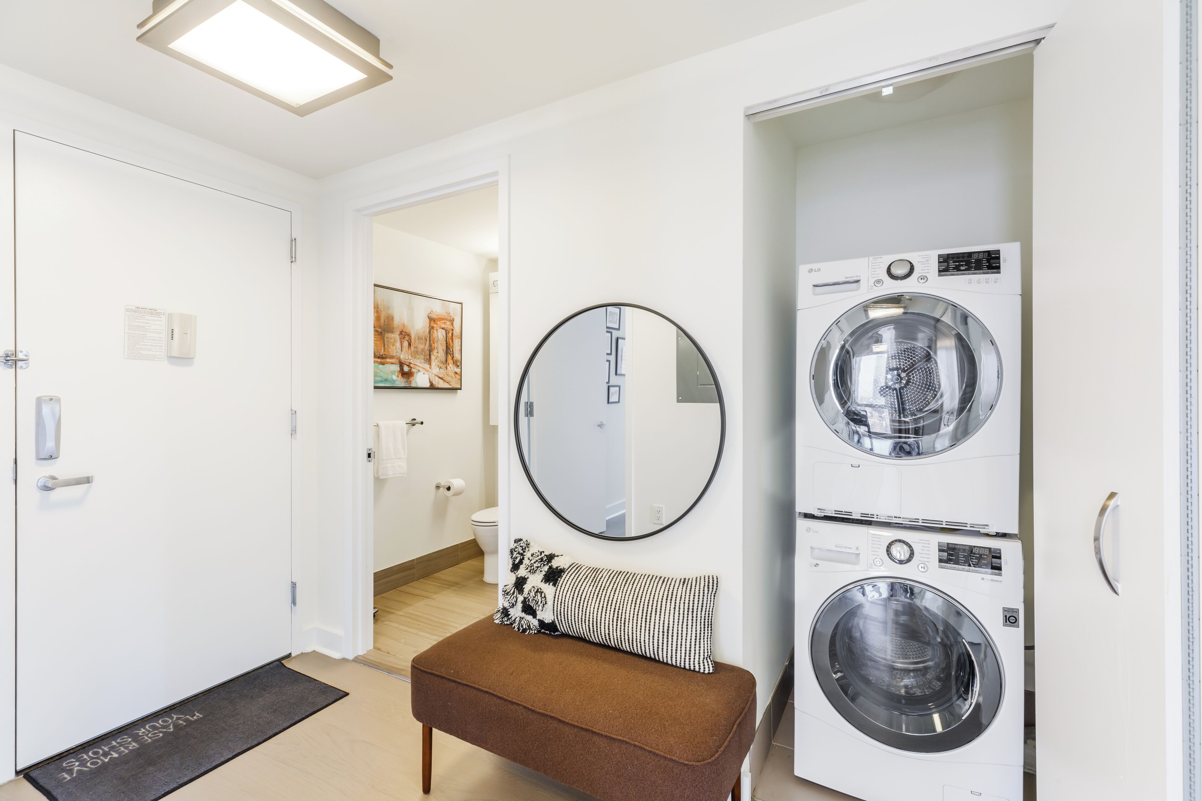 A laundry room with two stacked washing machines, a bench, a mirror, and a door leading to a bathroom.
