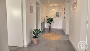 Een gang met witte muren, grijze vloer en twee potplanten. Er zijn twee hanglampen en een geweven vloerkleed.