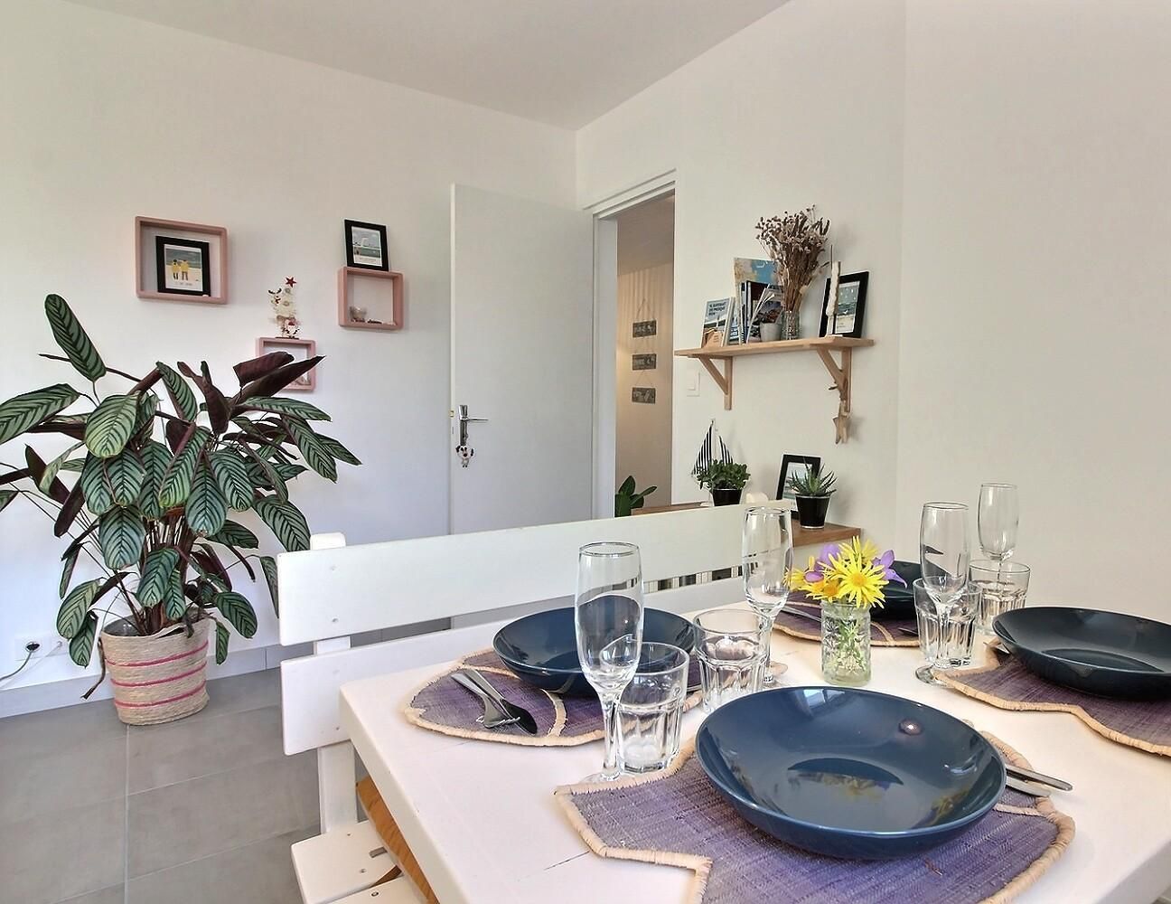 Un coin repas avec une table dressée pour quatre personnes, comprenant des assiettes, des verres et des couverts. On aperçoit une plante en pot sur la gauche et une étagère avec des objets décoratifs à l’arrière-plan.