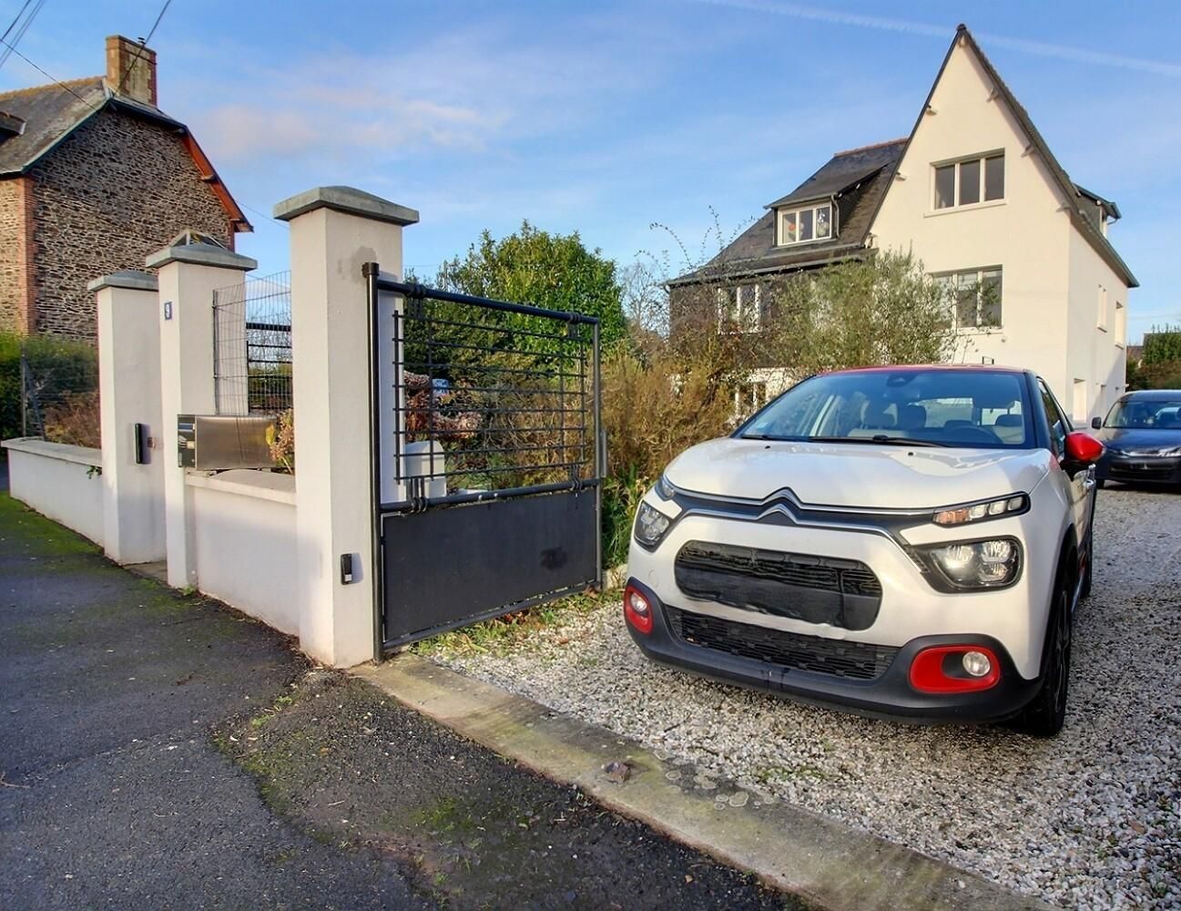 A white car parked in a driveway with a gated entrance, a house in the background, and a gravel path.