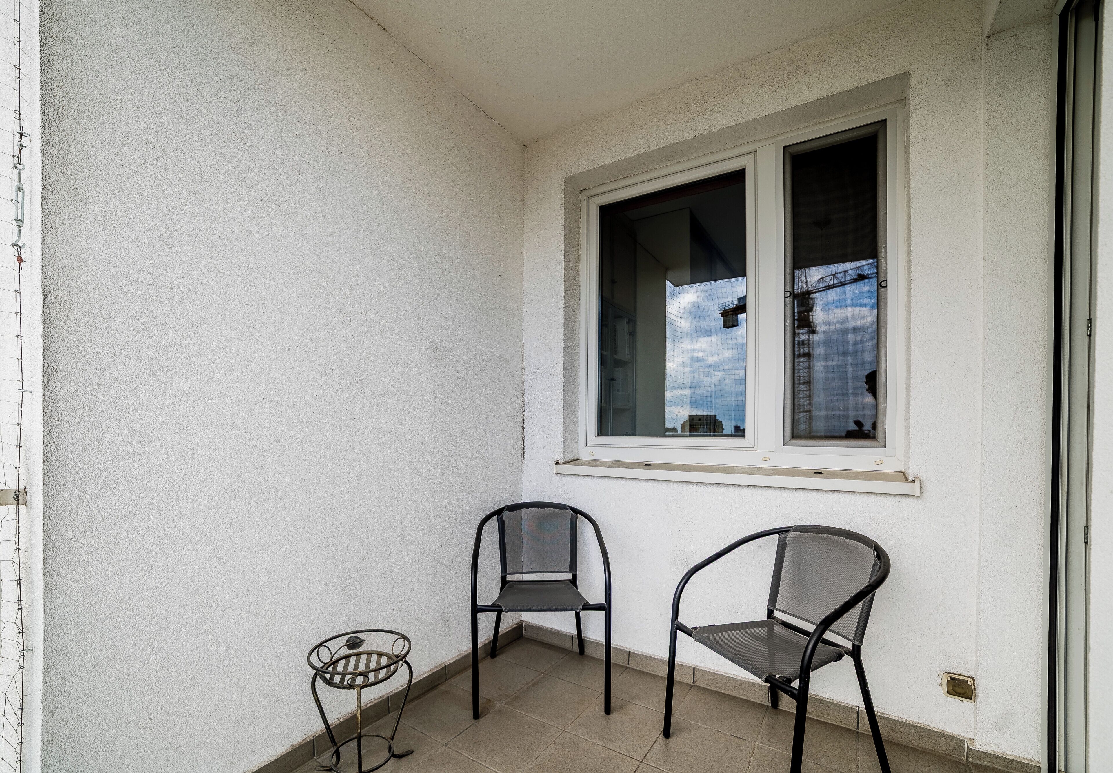 A balcony with two chairs and a small table, a window with a view of buildings and a blue sky, and a white wall.