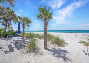 A beach with palm trees, a blue umbrella, and a concrete table. - Beautiful 2 BR Condo With Private Beach Access Sunset On The Gulf (Longboat Key)