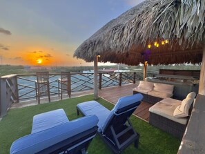 An outdoor deck with lounge chairs and a thatched roof structure, overlooking a body of water.