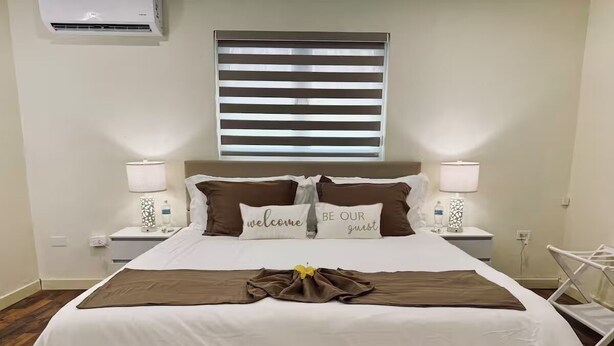 A neatly made bed with a brown throw, two pillows with "welcome" and "be our guest" text, and a bedside table with a lamp and a bottle.
