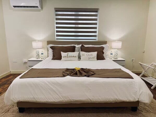 A neatly made bed with a brown throw, two pillows with "welcome" and "be our guest" text, and a bedside table with a lamp and a bottle.