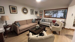 A living room with a beige sofa, a white armchair, and a coffee table. There are framed pictures on the wall and a window with a view of trees.