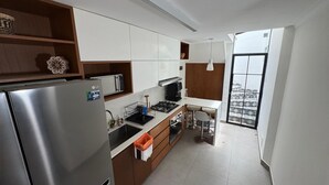 A modern kitchen with a stainless steel refrigerator, a microwave, and a gas stove. There is a small dining area with two chairs and a table. The kitchen has wooden cabinets and a large window providing natural light.