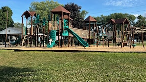 Ein Spielplatz mit verschiedenen Spielgeräten, darunter Rutschen und Kletterbereichen, umgeben von einer Rasenfläche und Bäumen.