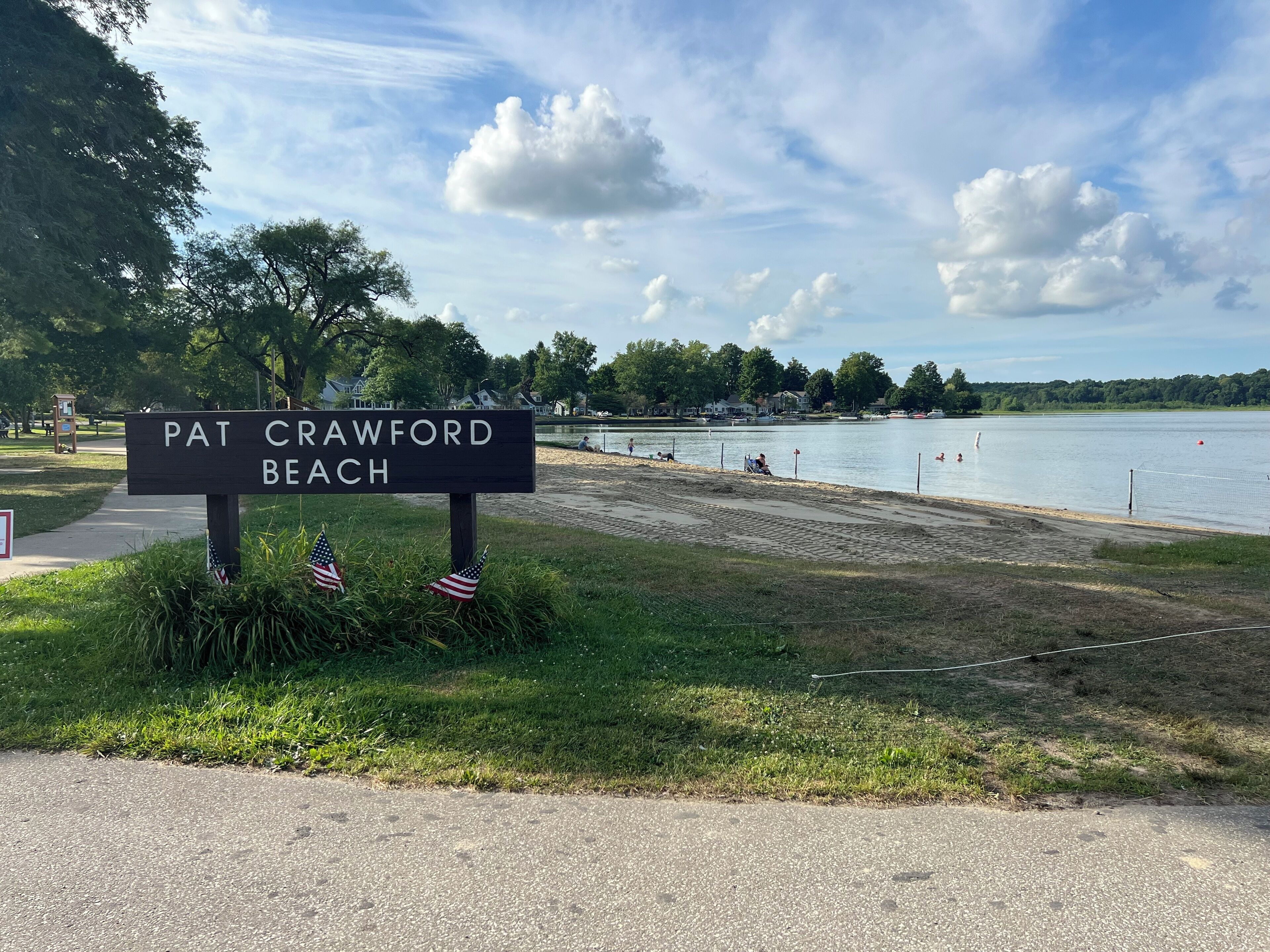 A beach sign with American flags, a lake, and houses in the background.