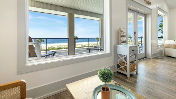 Ett modernt vardagsrum med ett stort fönster med utsikt över en strand, ett litet bord med en krukväxt och en vit soffa.
