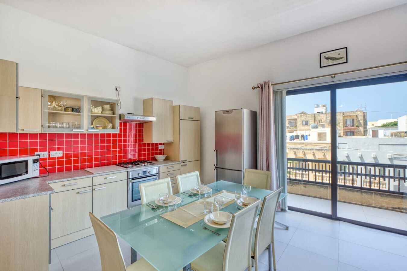 A modern kitchen with a red-tiled backsplash, a dining area with a glass table, and a balcony with a view of buildings.