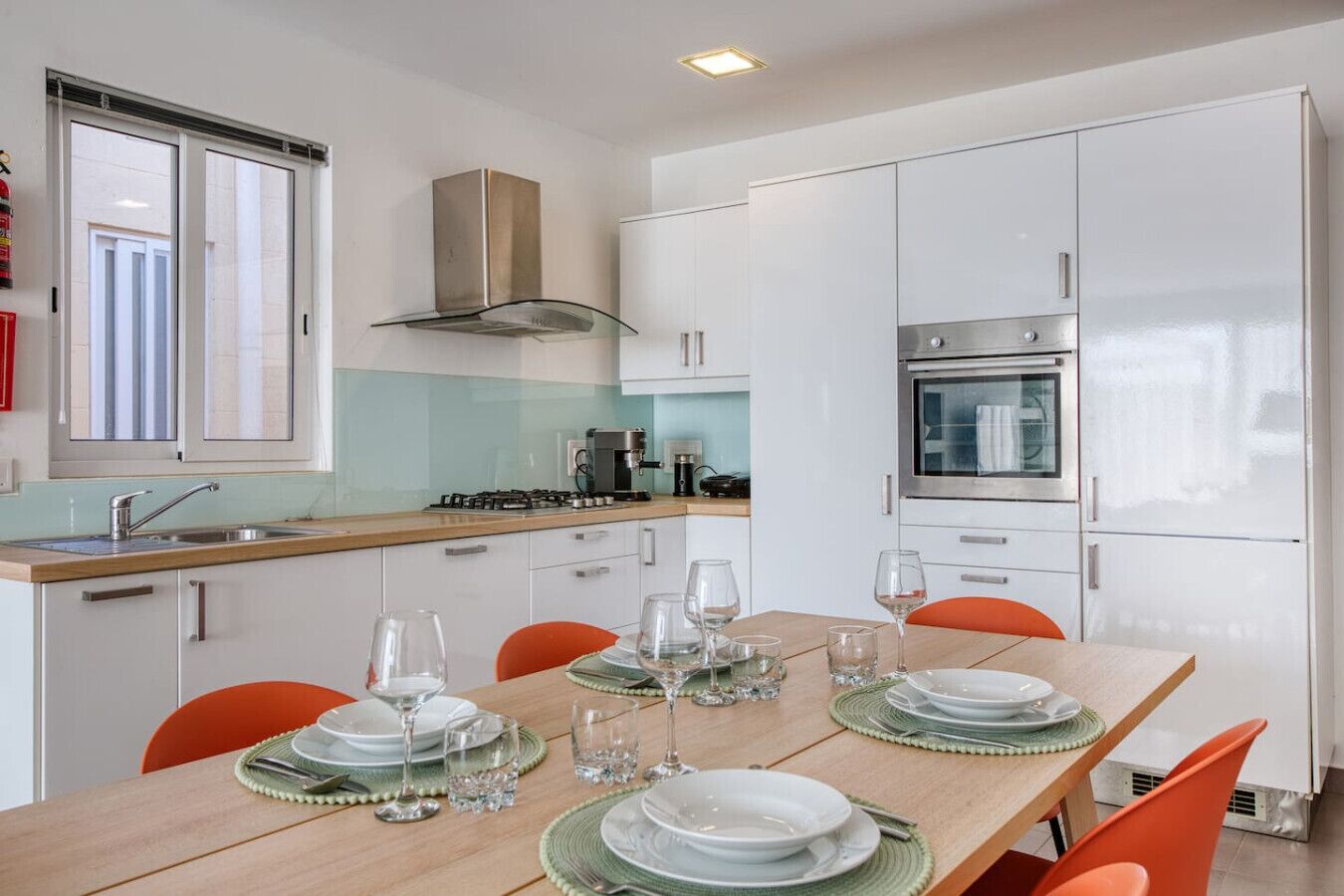 Une cuisine moderne avec des meubles blancs, un four en acier inoxydable et une cuisinière à gaz. On y trouve une table à manger mise avec des assiettes, des verres et des couverts.