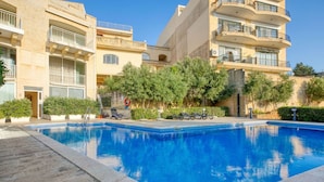 Μια πισίνα που περιβάλλεται από κτίρια με μπαλκόνια, δέντρα και έναν καθαρό ουρανό.