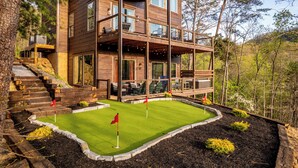 A multi-story wooden cabin with a deck, a small lawn with a flag, and a forested backdrop.