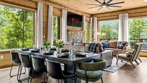 A spacious living area with a stone fireplace, a large TV screen displaying 'NETFLIX', and a view of the forest through large windows.