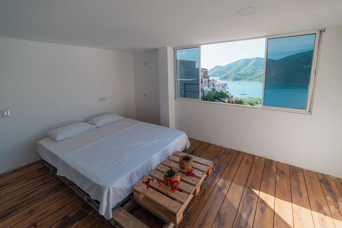 Un dormitorio con cama, piso de madera y un ventanal con vistas al agua y colinas.