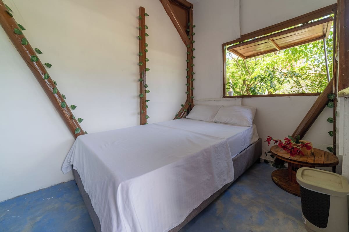 Un dormitorio con una cama, un ventanal con vistas a la vegetación y una mesita con una planta.