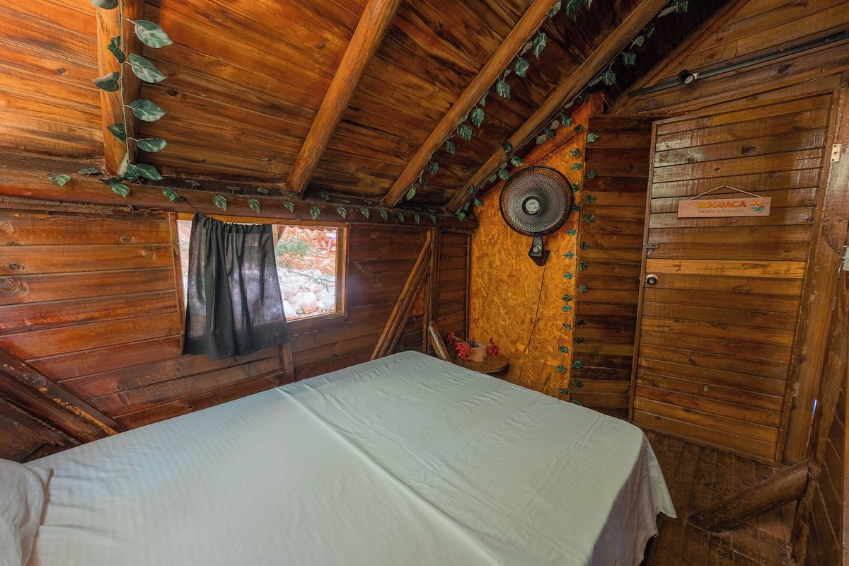 Habitación de cabaña de madera con una cama, una ventana con cortina, un ventilador en la pared y una puerta con un cartel.