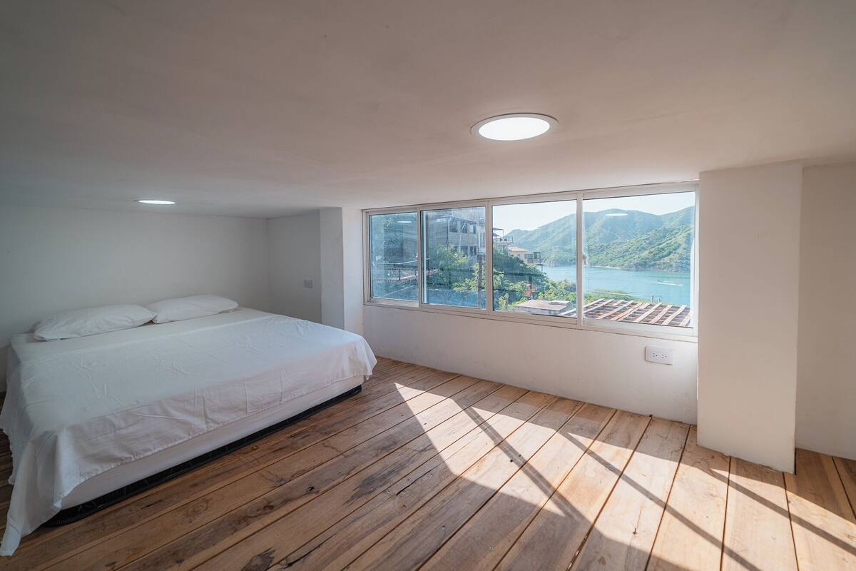 Un dormitorio con piso de madera, cama con ropa blanca, ventana con vista a un lago y colinas, y una lámpara de techo.