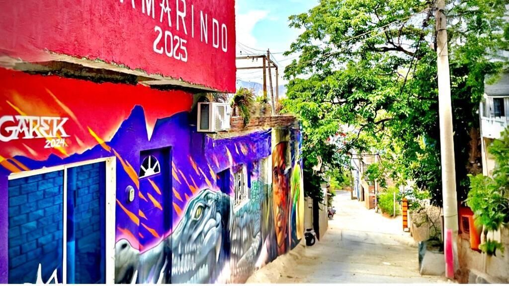 A narrow street with colorful murals on a building, a red banner with 'GARISK 2025', and a clear sky.