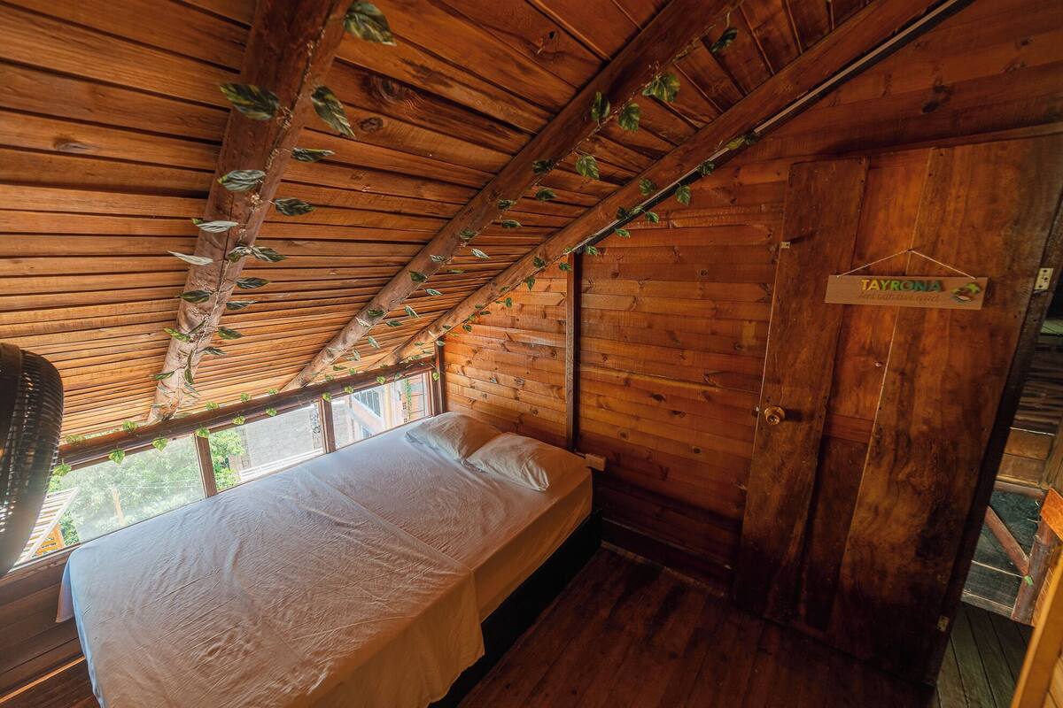 Una habitación tipo cabaña de madera con una cama, un ventanal con vistas y una puerta con la leyenda 'TAYRONA'.