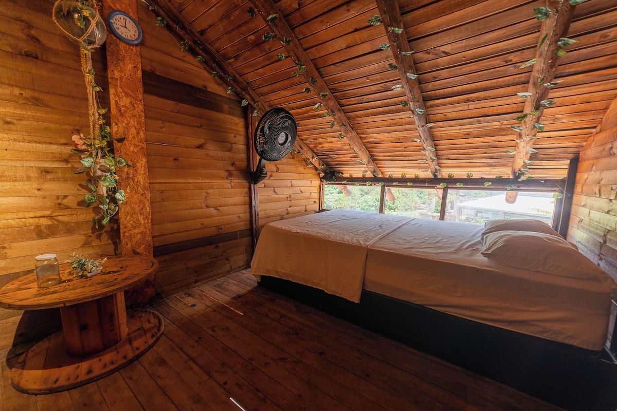 Una habitación cálida tipo cabaña con techo de madera, una cama, una mesita con un tarro de vidrio y un reloj de pared.