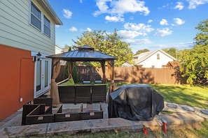 Ein Garten mit überdachter Terrasse, Sitzgelegenheiten im Freien und einem Grill.
