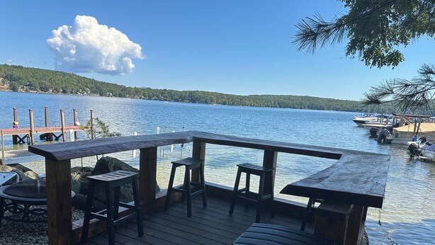 Una terraza de madera con un banco y una mesa, con vista a un lago tranquilo con barcos y un muelle al fondo.
