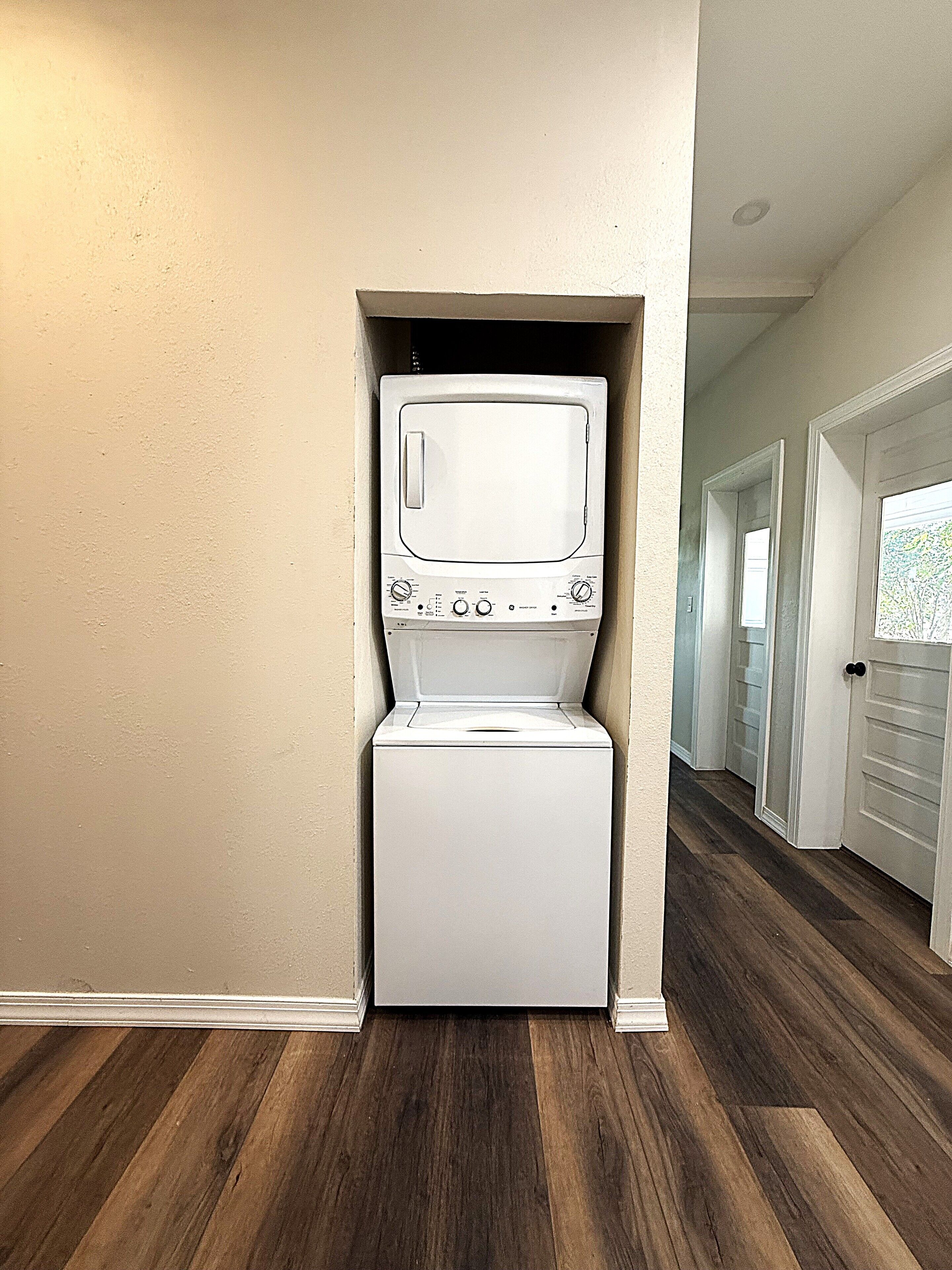 A laundry room with a front-loading washer and dryer, wooden flooring, and a hallway leading to other rooms.