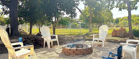 A backyard with a fire pit, white plastic chairs, and a blue folding chair.