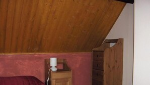Ein Schlafzimmer mit geneigter Decke, einem Holzschrank, einem Nachttisch mit Lampe und einer roten Bettdecke.