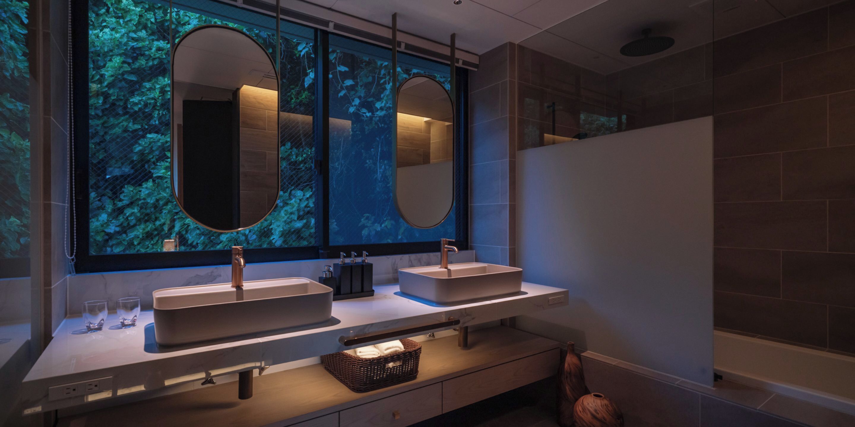 Un baño moderno con dos lavamanos, un espejo grande y un ventanal con vistas a la vegetación.