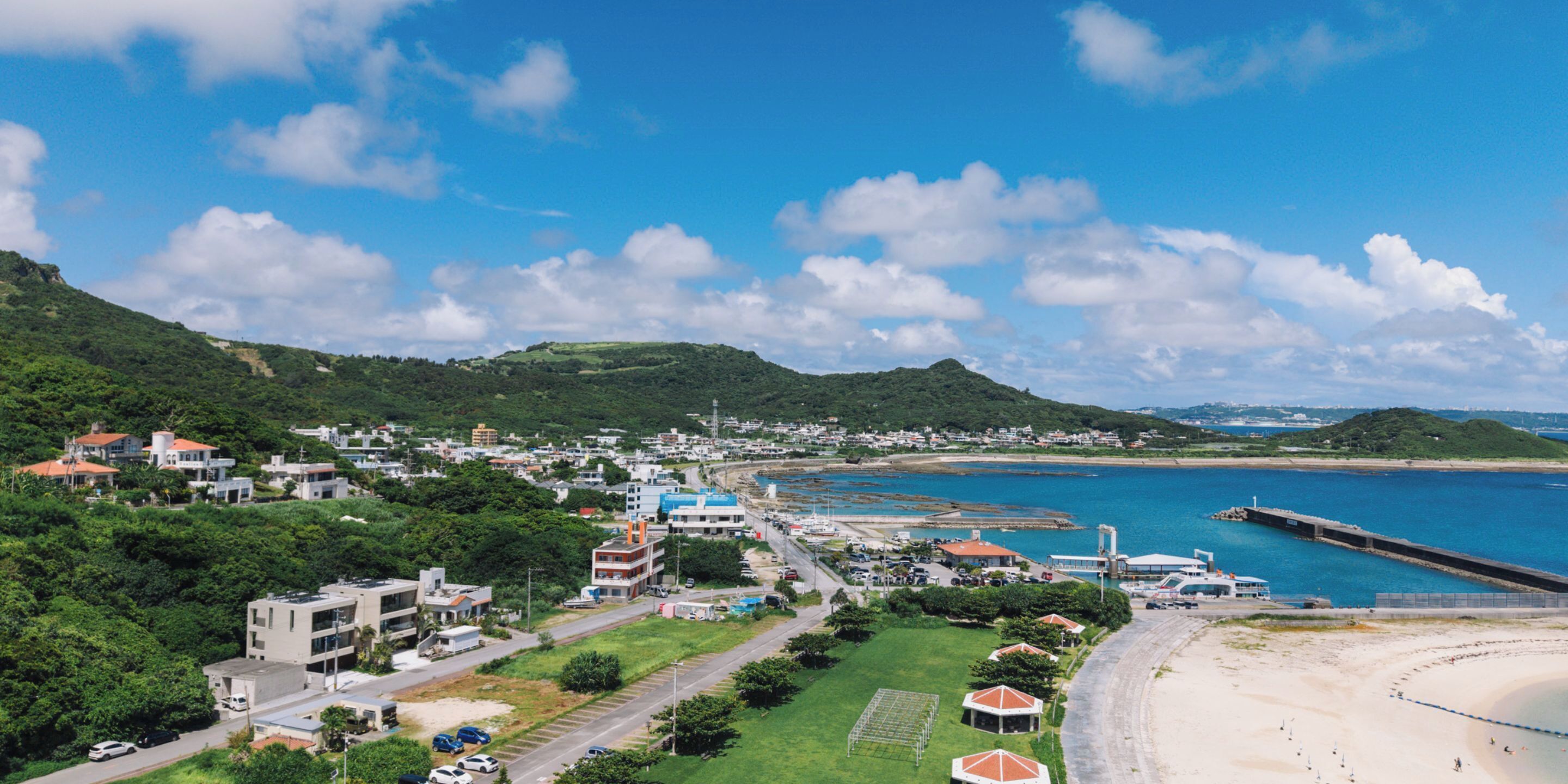An aerial view of a coastal town with a beach, green hills, and a mix of residential and commercial buildings.