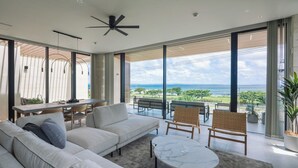 A modern living room with a large sofa, a dining table, and chairs. There is a view of the ocean through large windows. - CELEVI NANJO (Nanjo)