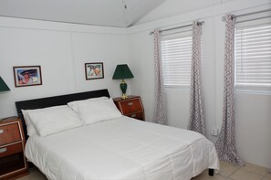 Una cama bien hecha con ropa blanca, una mesita de noche de madera con una lámpara, y dos cuadros en la pared.