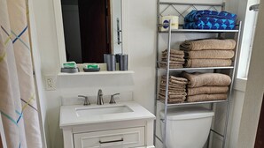 Un baño con lavamanos blanco, espejo e inodoro. Hay repisas con toallas dobladas y una cortina de ducha.