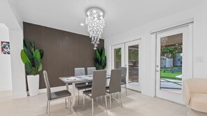 A modern dining area with a chandelier, grey upholstered chairs, and a large potted plant. - Tropical 4BR Villa with Pool, BBQ & Mini Golf Fun (Miami)