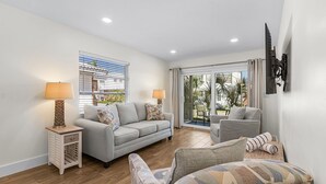 A living room with a grey sofa, a patterned armchair, a small side table with a lamp, and a sliding glass door leading to an outdoor area. - Unleash Island Bliss - Aloha Kai #34 & #37 - Roelens (Siesta Key)
